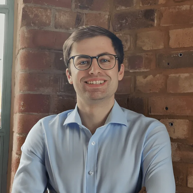 A young man wearing glasses and a light blue shirt is smiling while standing in front of a brick wall with crossed arms, embodying the spirit of sustainability at Cority. Natural light comes from a window on the left.