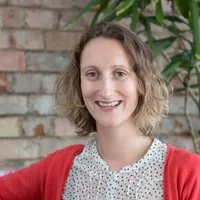 A woman with wavy, shoulder-length light brown hair smiles at the camera. She wears a white blouse with black polka dots and a red cardigan, standing by a brick wall and leafy plant—reflecting the spirit of sustainability at Cority.