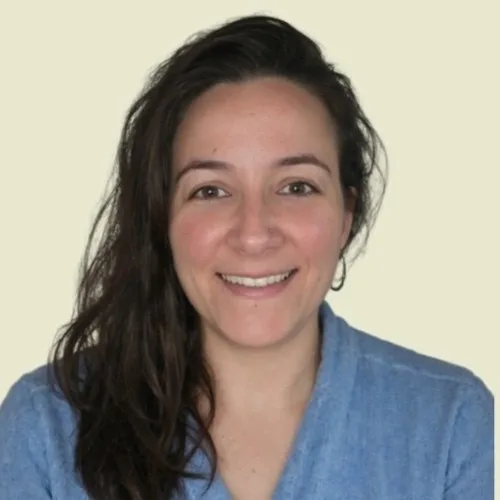 A woman with long dark hair, wearing a blue top, is smiling at the camera against a plain light background, reflecting the approachable spirit of Sustainability at Cority.