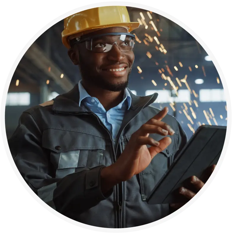 A smiling industrial worker wearing a yellow hard hat, safety glasses, and a protective jacket uses a tablet, with bright sparks flying in the background of a factory setting.