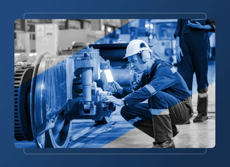 A worker wearing a helmet and ear protection inspects a train wheel assembly with a flashlight in an industrial setting, similar to those in the aerospace industry, while another person stands nearby with a clipboard.