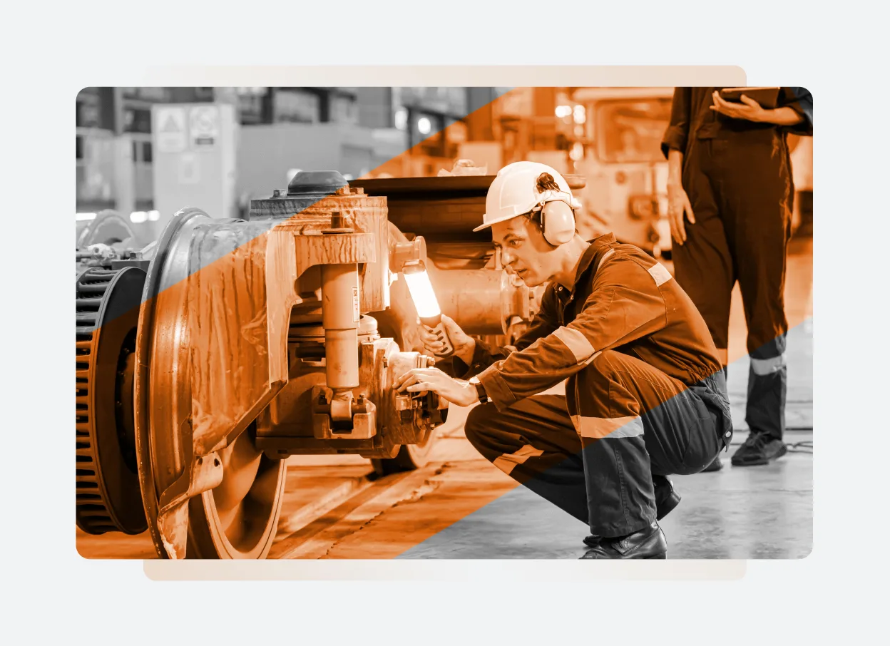 A worker wearing a hard hat and ear protection inspects and repairs machinery on a large industrial vehicle in a factory setting, showcasing how Solutions Main streamlines maintenance. Another person stands in the background, partially visible.