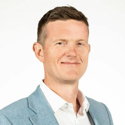 A man with short brown hair wearing a light blue blazer and white shirt, smiling slightly while standing against a plain white background, embodies the spirit of sustainability at Cority.