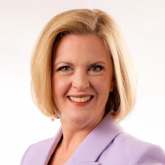 A woman with short blonde hair is smiling at the camera. She is wearing a light purple blazer and is posed against a plain white background.