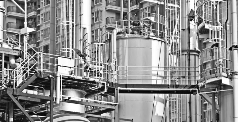A black-and-white photo of an industrial structure with metal pipes, tanks, and platforms, set against the background of a high-rise residential building with many windows.