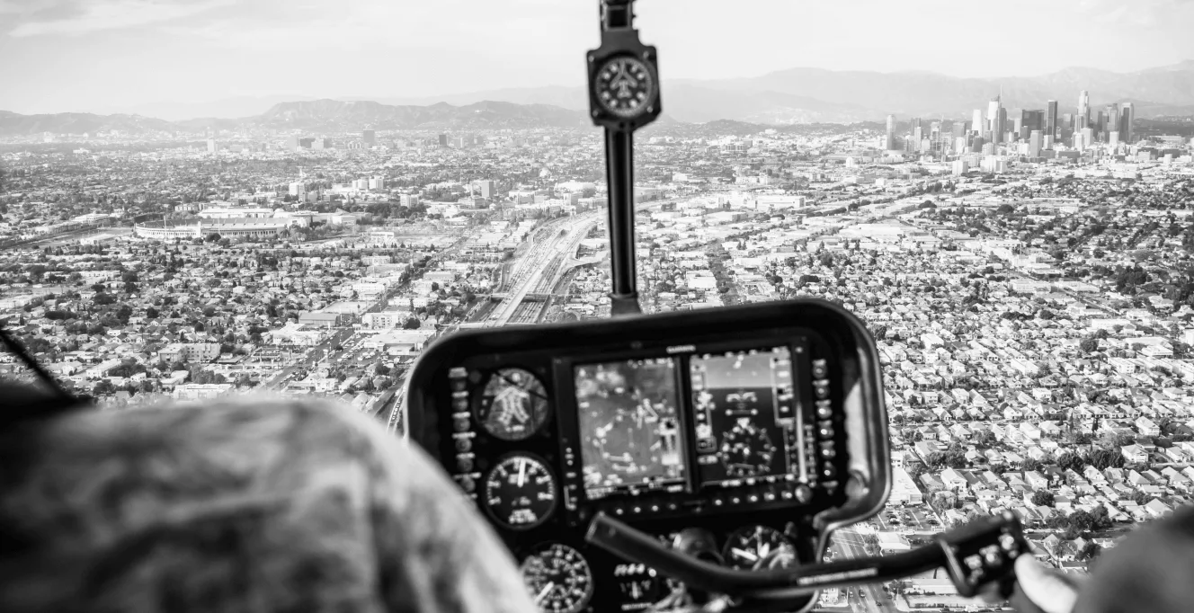 A black-and-white aerial view of a city, seen from inside a helicopter cockpit with visible controls and screens, while distant skyscrapers and mountains are in the background.