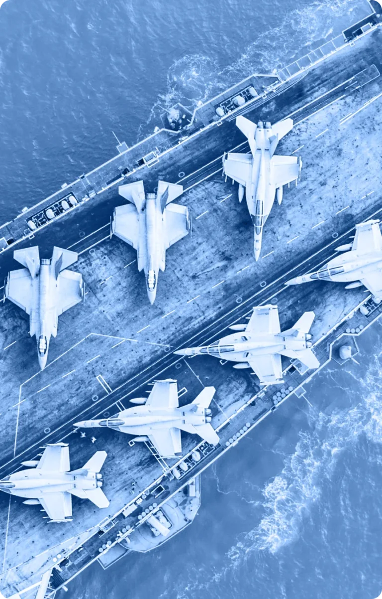 Aerial view of six fighter jets arranged on the deck of an aircraft carrier at sea, with waves visible around the ship. The image has a blue color filter, suitable for use in an Industry Tab Section.