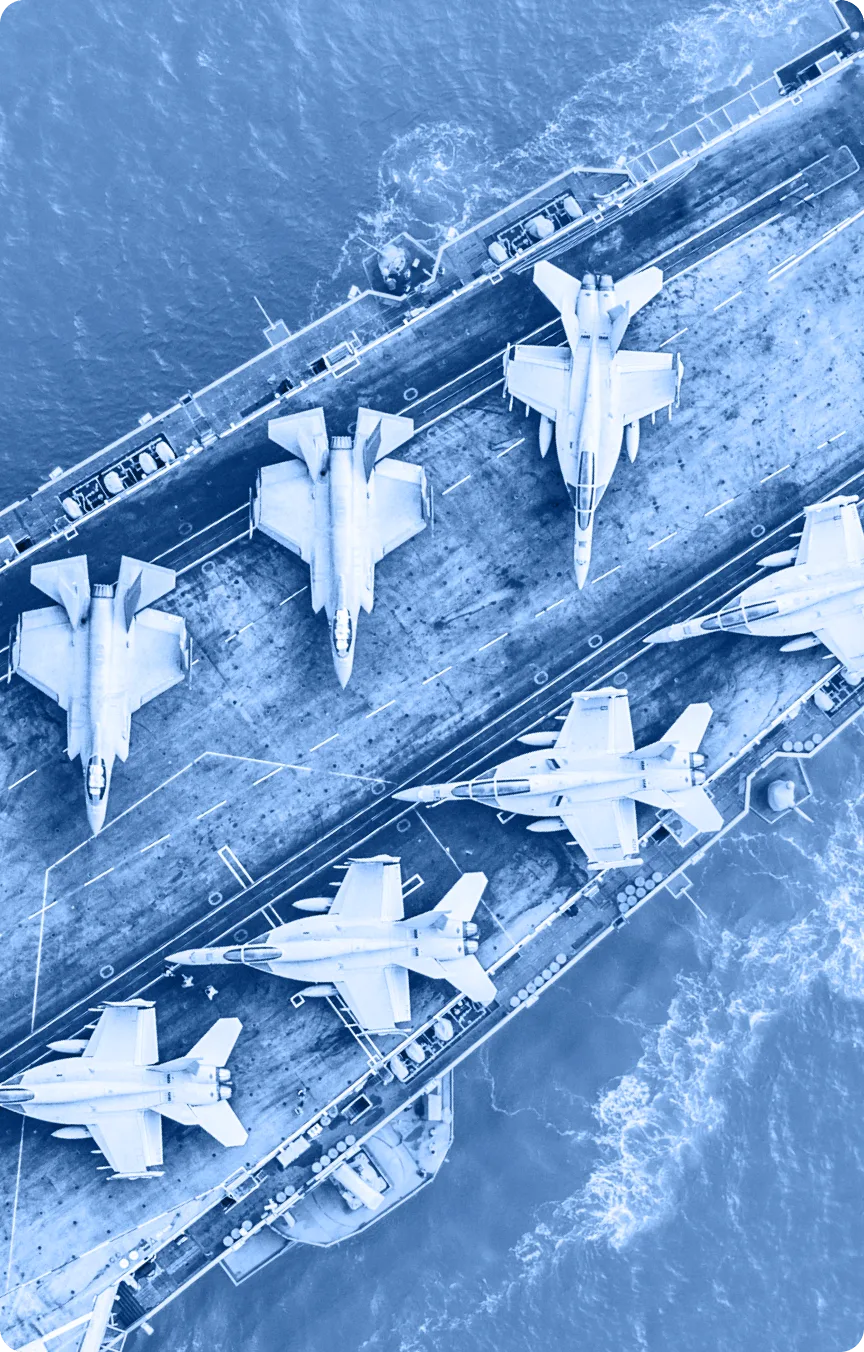Aerial view of six fighter jets arranged on the deck of an aircraft carrier at sea, with waves visible around the ship. The image has a blue color filter, suitable for use in an Industry Tab Section.