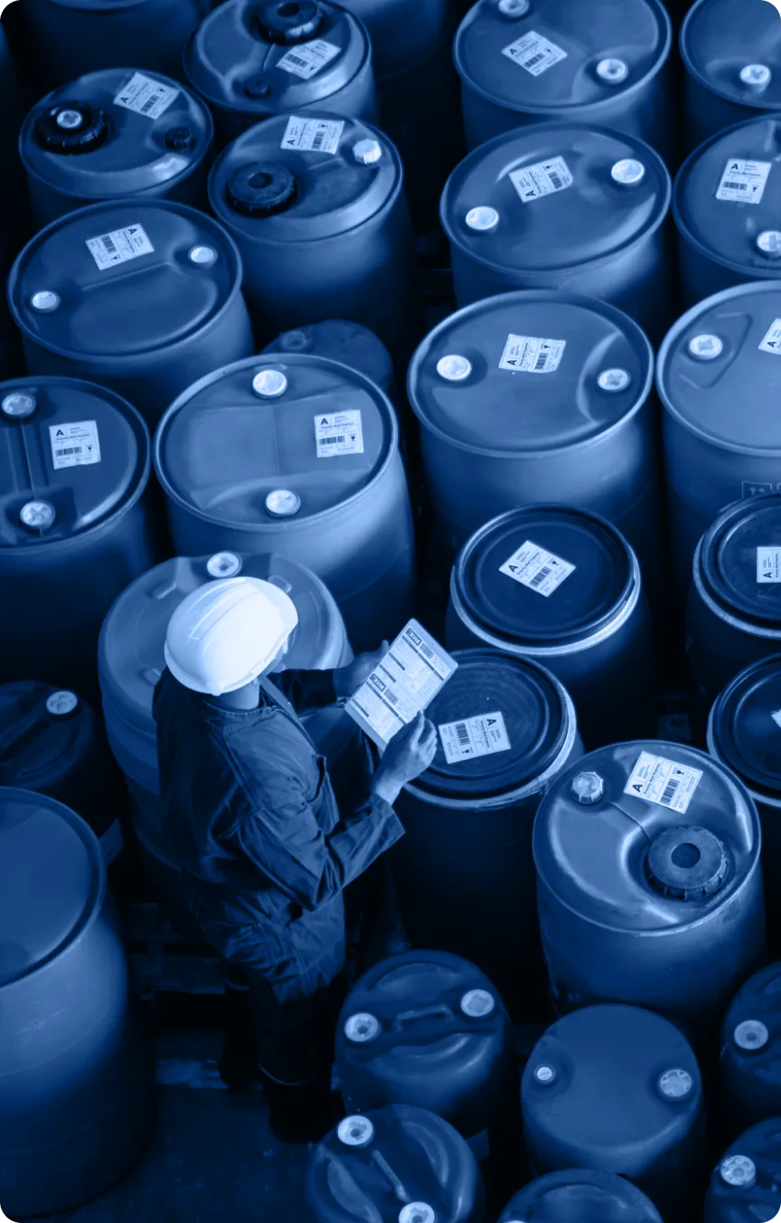 A person wearing a white hard hat and dark clothing checks a clipboard while standing among rows of large, labeled industrial barrels arranged in a storage area, highlighting the importance of safety and organization in the industry.