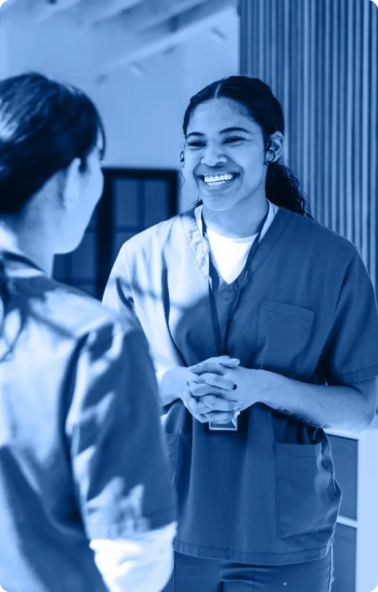 Two healthcare workers in scrubs stand facing each other indoors. One woman is smiling warmly and clasping her hands while speaking to the other, who has her back to the camera, highlighting collaboration in the Industry Section.