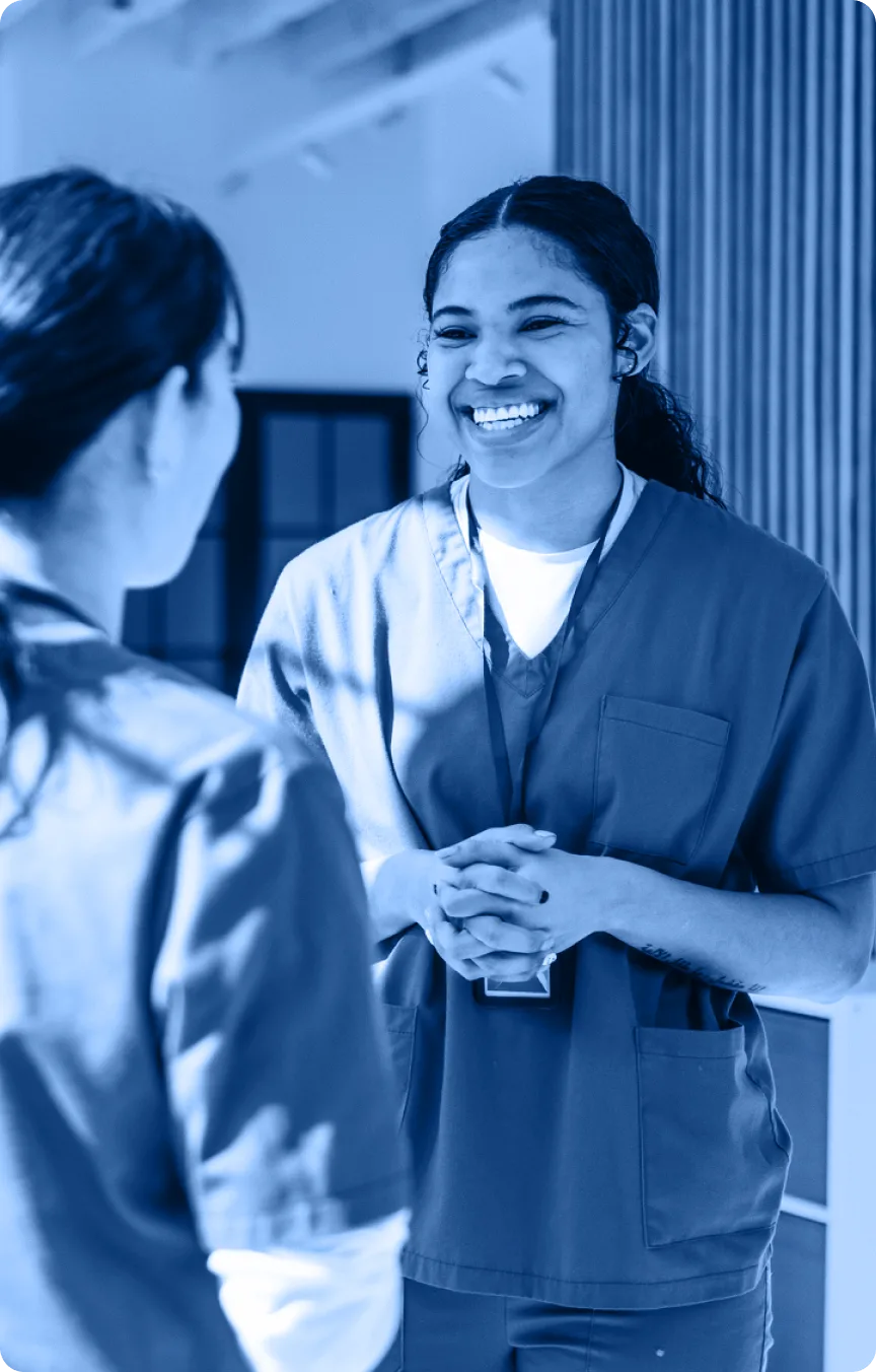 Two healthcare workers in scrubs stand facing each other indoors. One woman is smiling warmly and clasping her hands while speaking to the other, who has her back to the camera, highlighting collaboration in the Industry Section.