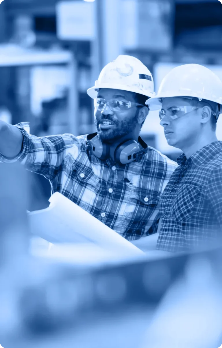 Two construction workers in hard hats, safety glasses, and plaid shirts discuss plans at a worksite. One holds blueprints and points ahead as they focus on the project, ready to be featured in the Industry Tab Section.