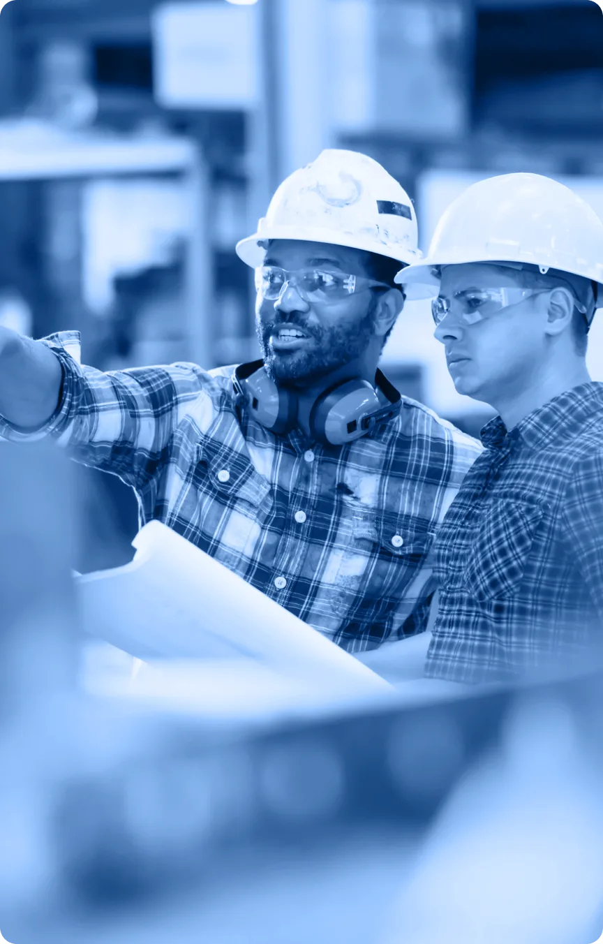 Two construction workers in hard hats, safety glasses, and plaid shirts discuss plans at a worksite. One holds blueprints and points ahead as they focus on the project, ready to be featured in the Industry Tab Section.