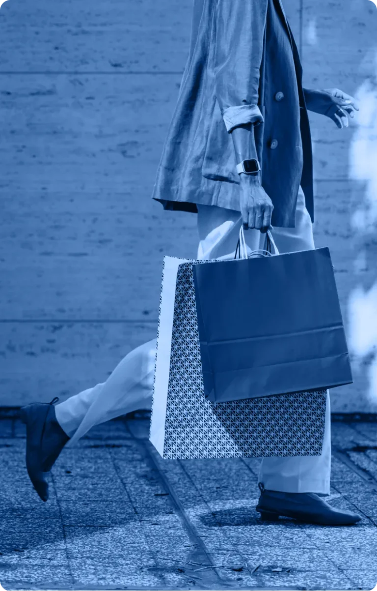 A person wearing a blazer, pants, loafers, and a smartwatch walks outdoors with two large shopping bags. The image, reminiscent of an Industry Section visual, is enhanced by a striking blue color filter.