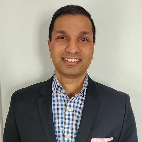 A man with short dark hair smiles at the camera. He is wearing a dark blazer over a blue and white checkered shirt, a light pink pocket square, reflecting the professionalism seen in Sustainability at Cority. The background is plain and light-colored.