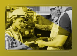 Two workers in safety gear discuss something on a digital tablet in an industrial setting, with machinery visible and a yellow overlay. They review data from the Safety Cloud, enhancing workplace safety and efficiency.
