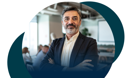 A confident middle-aged man with a beard, wearing a suit, stands with his arms crossed in a modern office setting. Blurred colleagues and minimalist decor are visible in the background.