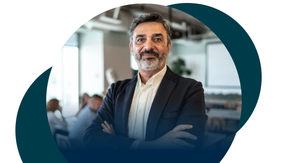 A confident middle-aged man with a beard, wearing a suit, stands with his arms crossed in a modern office setting. Blurred colleagues and minimalist decor are visible in the background.