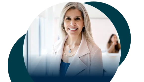 A woman with long, light hair, wearing a light-colored blazer and necklace, smiles at the camera in a bright office. Another person is blurred in the background. Circular blue graphic shapes overlay the image edges.