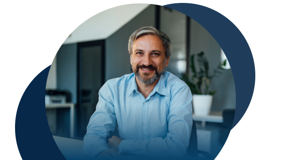 A man with gray hair and a beard, wearing a light blue shirt, smiles while sitting at a desk in a modern office with plants and glass doors in the background.