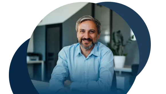 A man with gray hair and a beard, wearing a light blue shirt, smiles while sitting at a desk in a modern office with plants and glass doors in the background.