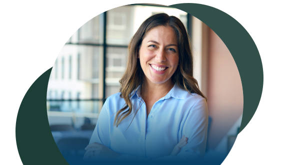 A woman with long brown hair, wearing a light blue blouse, smiles confidently with arms crossed in a bright, modern office setting.