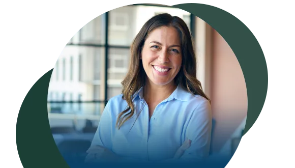 A woman with long brown hair, wearing a light blue blouse, smiles confidently with arms crossed in a bright, modern office setting.
