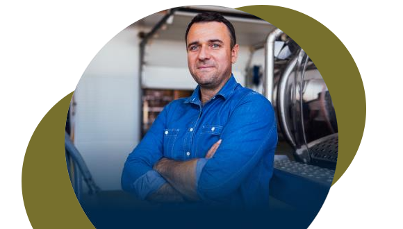 A man in a blue button-up shirt stands with arms crossed, smiling confidently in an industrial or workshop setting, with abstract circular shapes framing the image.