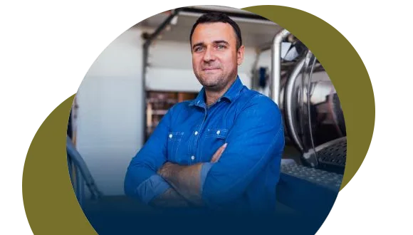 A man in a blue button-up shirt stands with arms crossed, smiling confidently in an industrial or workshop setting, with abstract circular shapes framing the image.