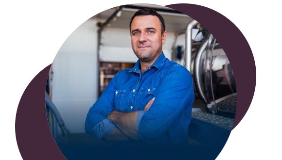 A man in a blue denim shirt stands with his arms crossed, smiling confidently. He is indoors, possibly in a workshop or industrial setting, with equipment visible in the background.