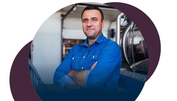 A man in a blue denim shirt stands with his arms crossed, smiling confidently. He is indoors, possibly in a workshop or industrial setting, with equipment visible in the background.