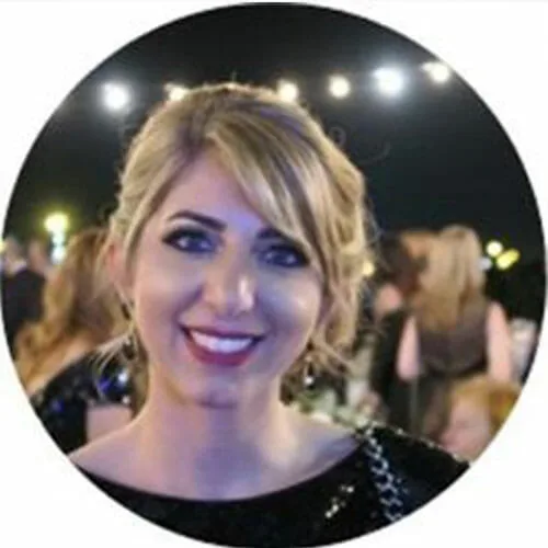 A woman with blonde hair and red lipstick smiles at the camera during an outdoor nighttime event celebrating sustainability at Cority, with blurred people and string lights glowing in the background.