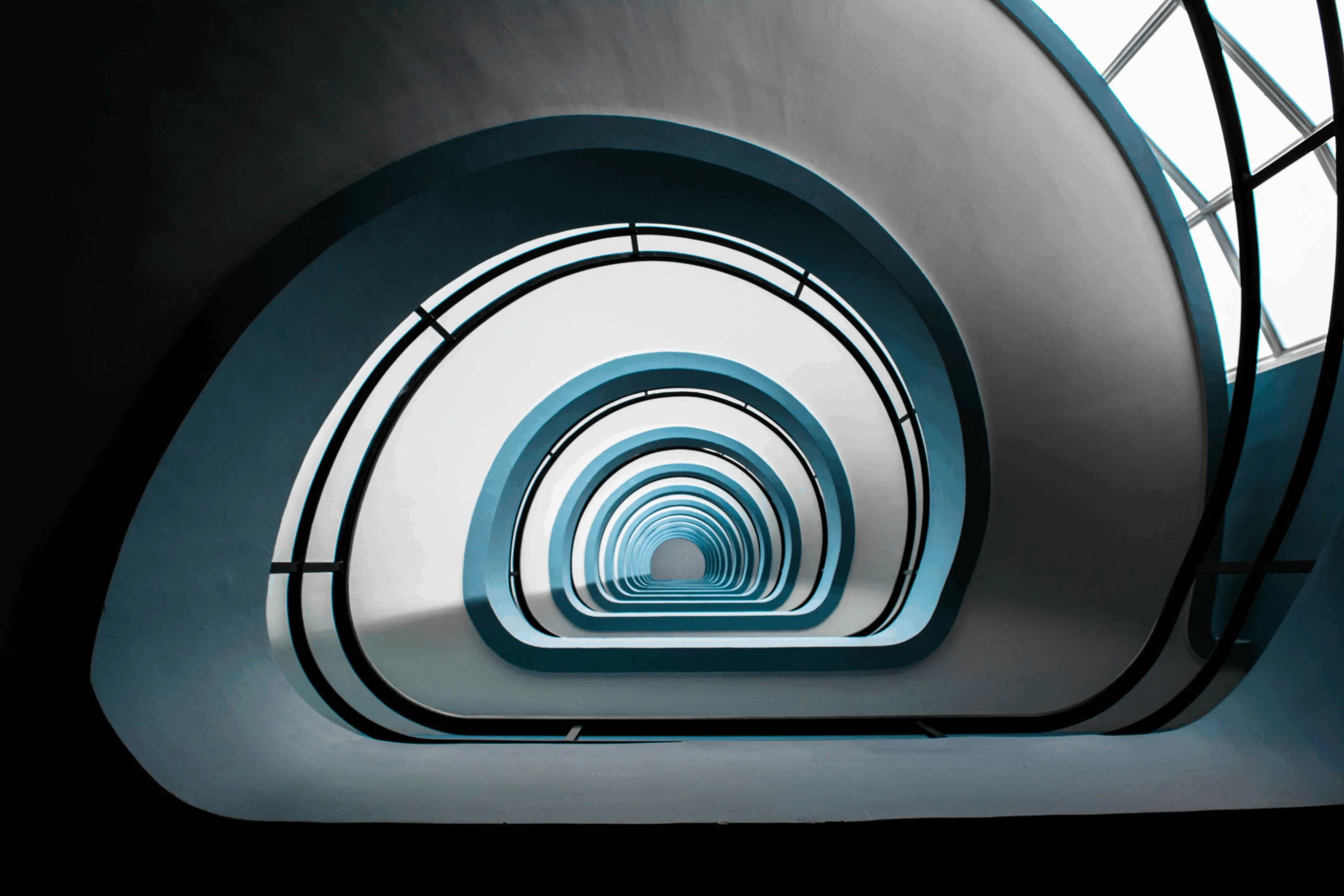 A view looking up through a spiral staircase with smooth white and blue curves, resembling the streamlined effect of ehs software consolidation, creating a symmetrical, tunnel-like pattern that leads the eye toward a bright light at the top.