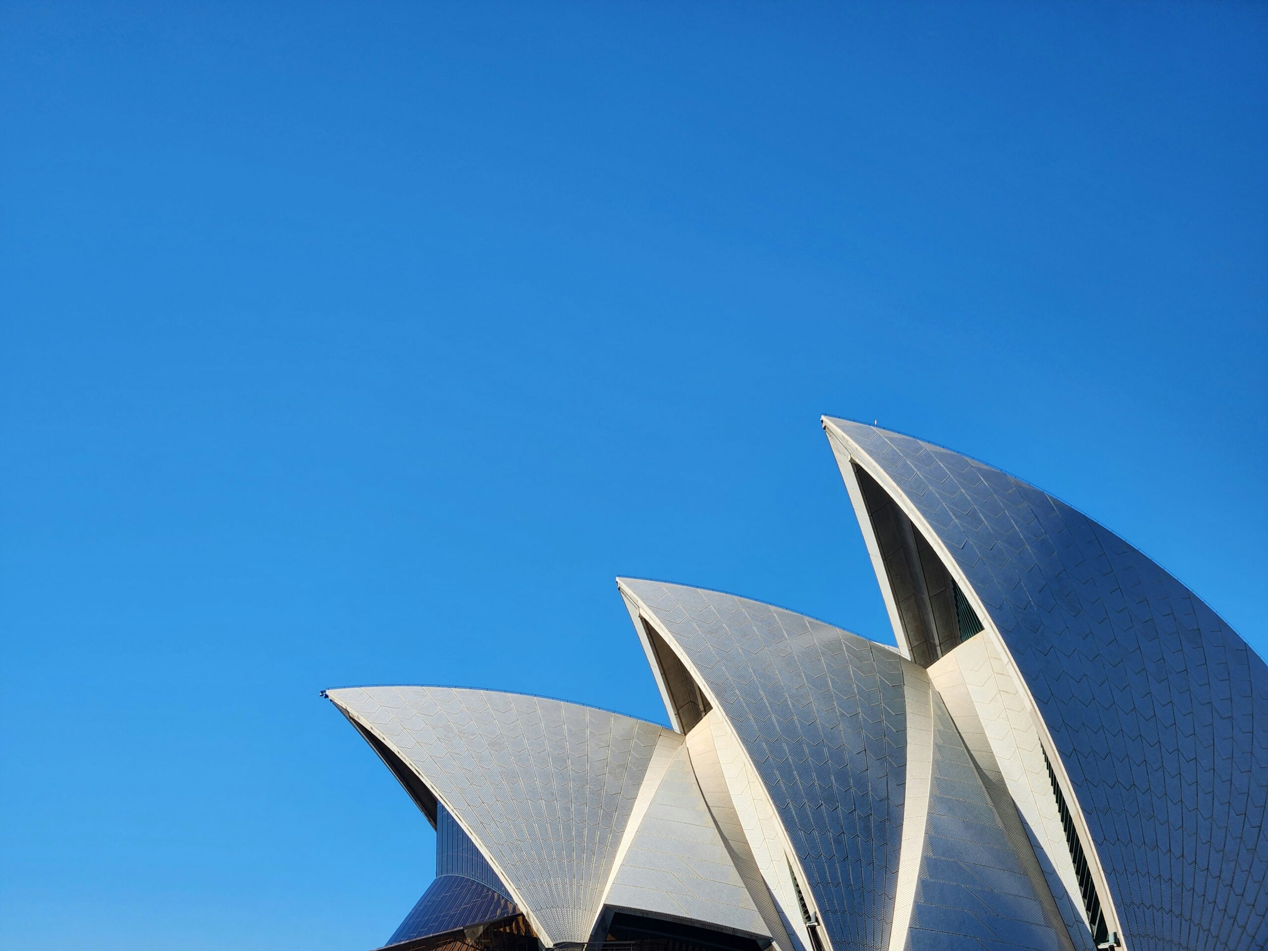 The white, sail-like roof structures of the Sydney Opera House stand out against a clear blue sky, with only the tops of the building visible in the image.