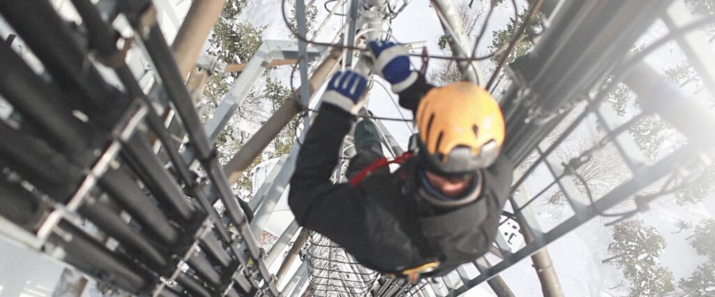 A worker wearing a yellow helmet and safety gear climbs a tall communications tower as part of digital incident management, gripping metal rungs beside large cables, with a snowy landscape visible below.