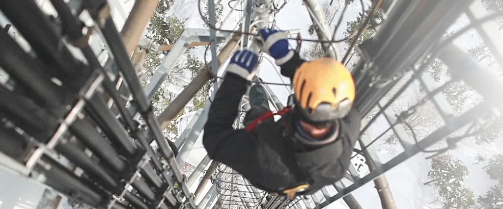 A worker wearing a yellow helmet and safety gear climbs a tall communications tower as part of digital incident management, gripping metal rungs beside large cables, with a snowy landscape visible below.