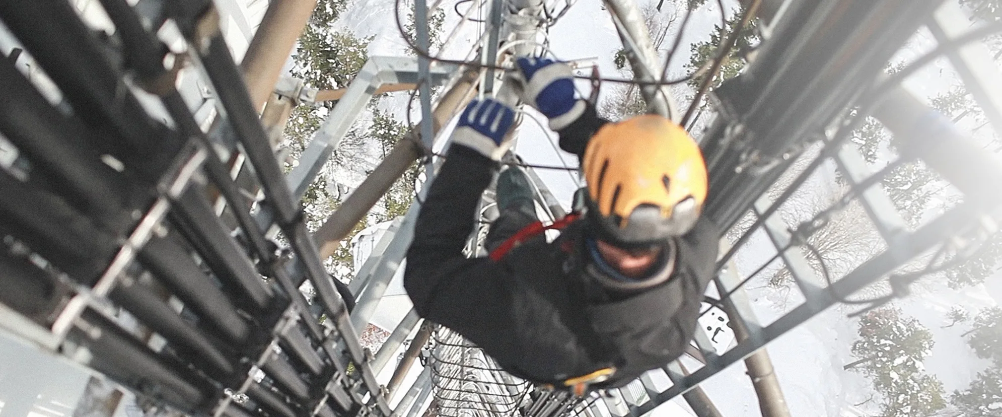 A worker wearing a yellow helmet and safety gear climbs a tall communications tower as part of digital incident management, gripping metal rungs beside large cables, with a snowy landscape visible below.