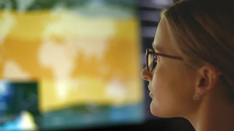 A woman wearing glasses looks at a large, blurred screen displaying a yellow-toned world map, suggesting data analysis or monitoring of leading safety indicators in a technology-driven environment.