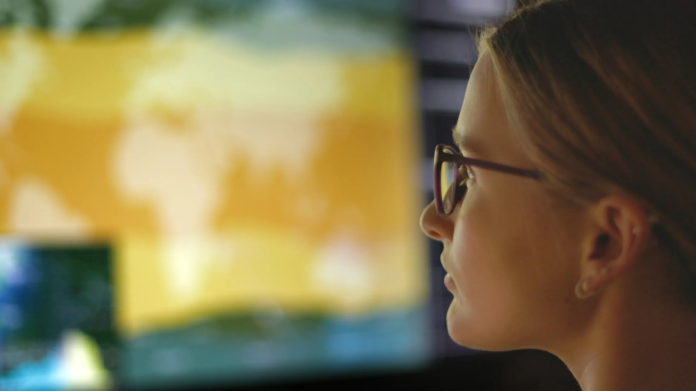 A woman wearing glasses looks at a large, blurred screen displaying a yellow-toned world map, suggesting data analysis or monitoring of leading safety indicators in a technology-driven environment.