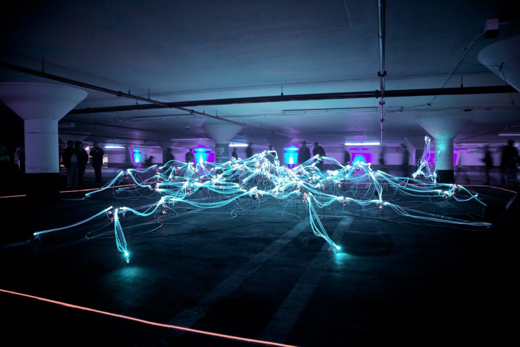 A dimly lit parking garage with glowing blue and purple neon light trails sprawling across the floor; several people stand in the background near illuminated pillars, discussing applied AI in EHS to enhance safety measures.