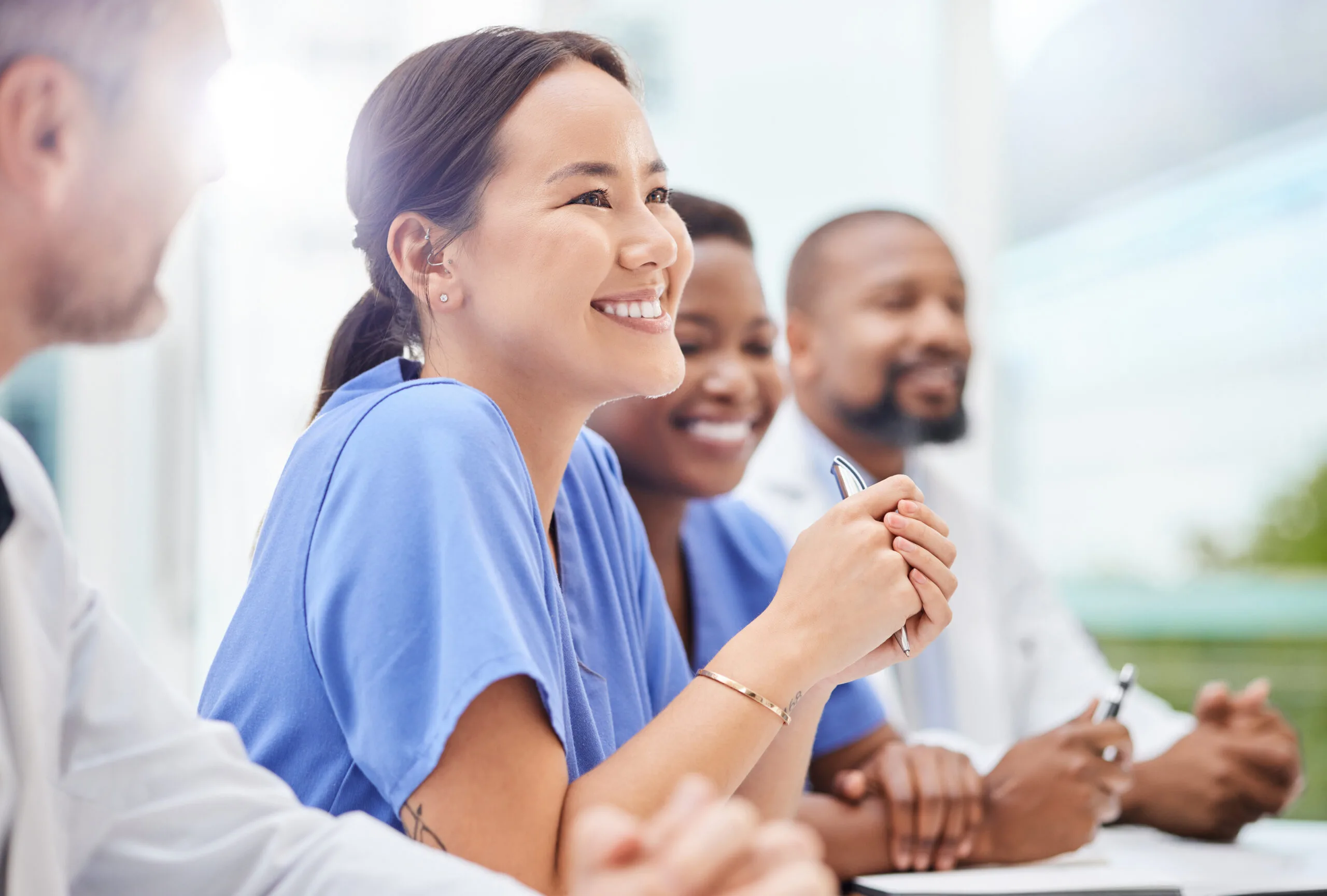 A group of four healthcare professionals in scrubs and lab coats sit together at a table, smiling and engaged, as they discuss the difference between health and safety. One woman in blue scrubs holds a pen and looks thoughtfully to the side.