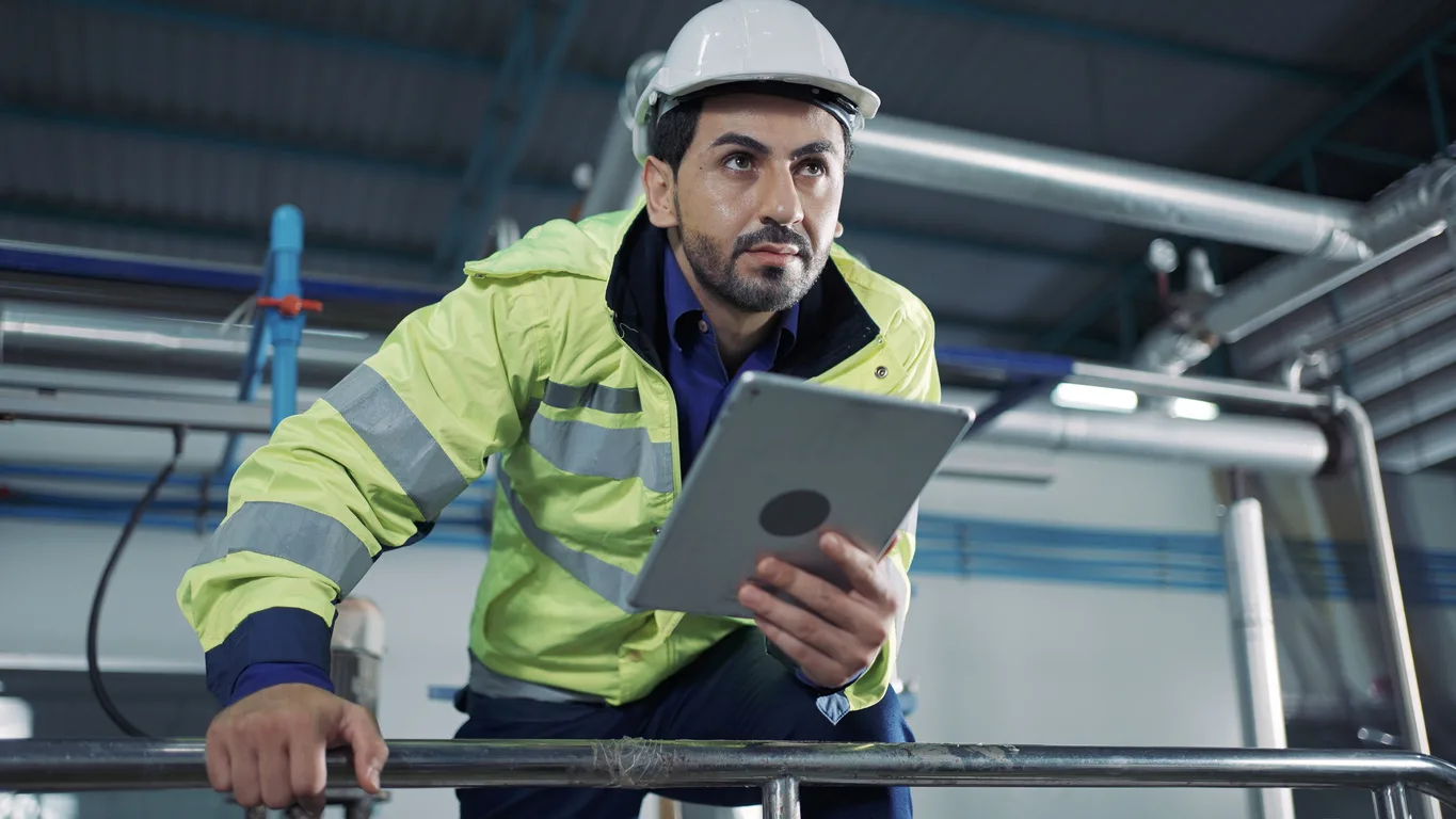 A man wearing a white hard hat and a high-visibility jacket holds a tablet while inspecting industrial equipment, utilizing ai for workplace safety in a factory or plant setting.