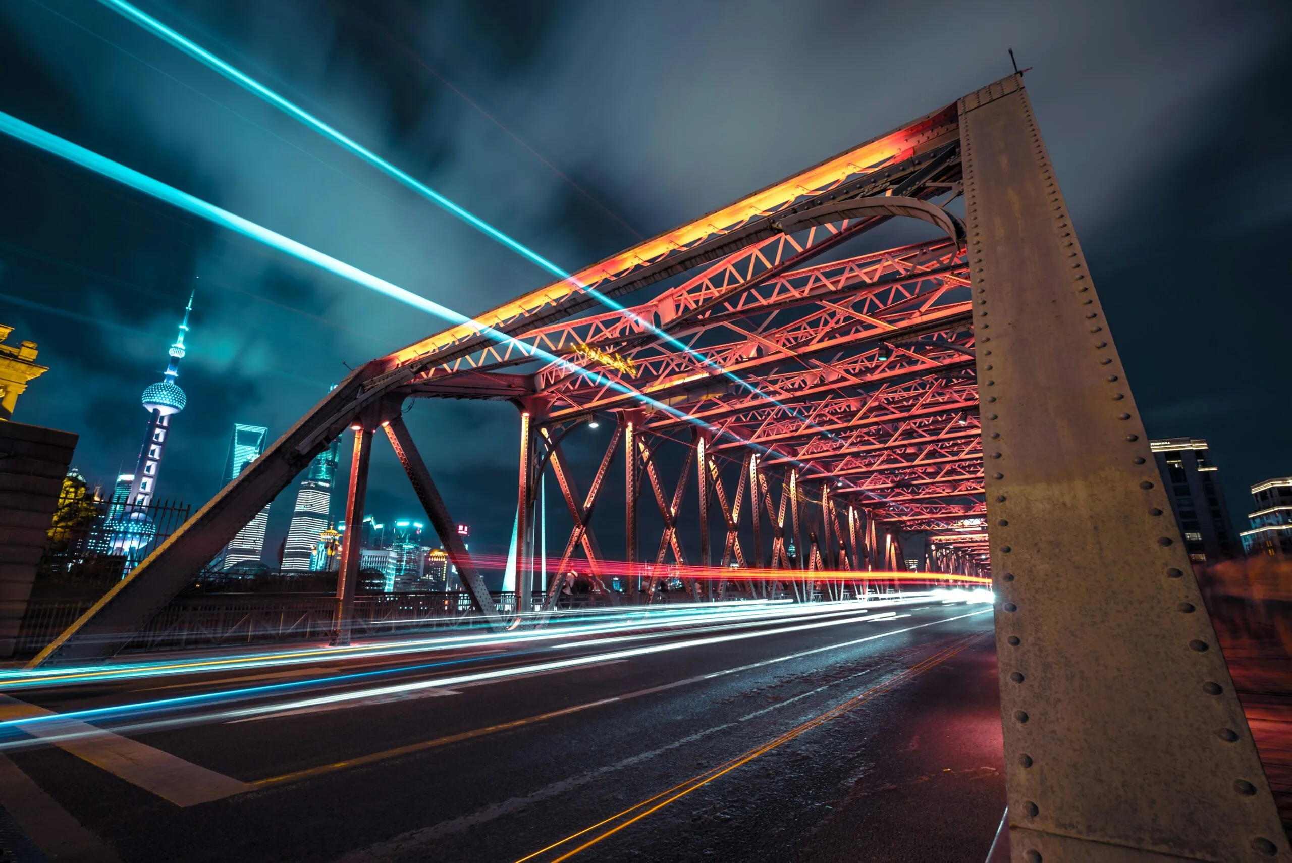 A steel bridge at night with red lights illuminating its structure. Light trails from moving vehicles create streaks across the road, while sustainability teams admire the city skyline and a tall, lit-up tower in the background.