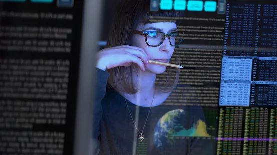 A woman wearing glasses looks thoughtfully at multiple computer screens displaying code and data charts, a pencil to her lips, suggesting deep concentration on automotive data security and TISAX certification requirements.