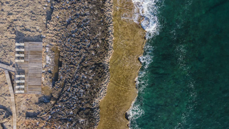 Aerial view of a rocky shoreline with clear blue-green ocean waves meeting a textured, tan rocky coast. A row of empty lounge chairs sits on a wooden deck, suggesting sustainability—ideal for showcasing scalable PFAS management solutions.