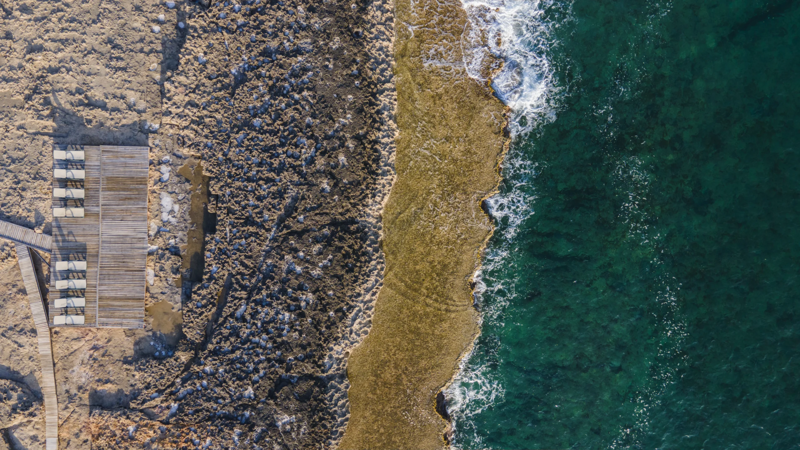 Aerial view of a rocky shoreline with clear blue-green ocean waves meeting a textured, tan rocky coast. A row of empty lounge chairs sits on a wooden deck, suggesting sustainability—ideal for showcasing scalable PFAS management solutions.